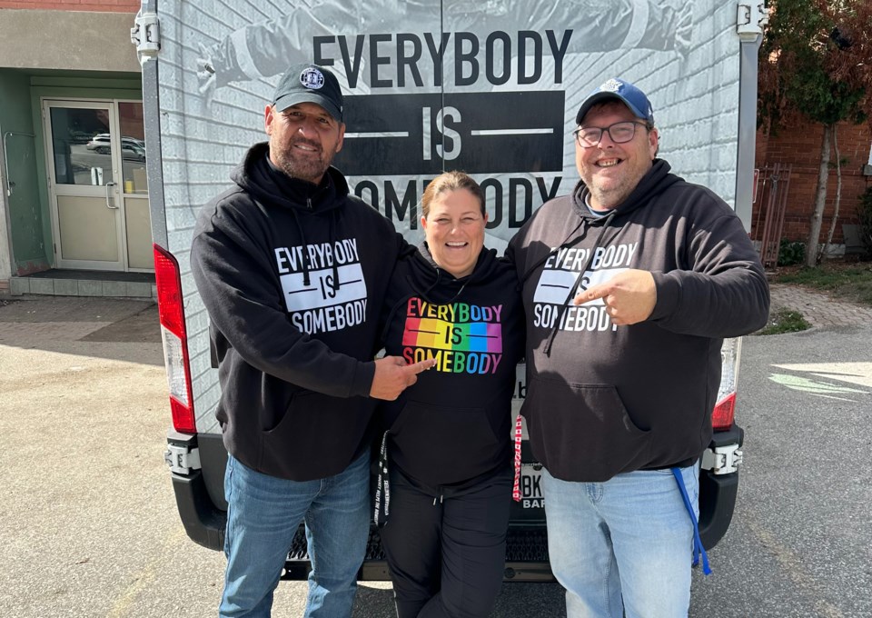 Three volunteers smile at the camera wearing sweatshirts printed with the slogan, "Everybody is Somebody"
