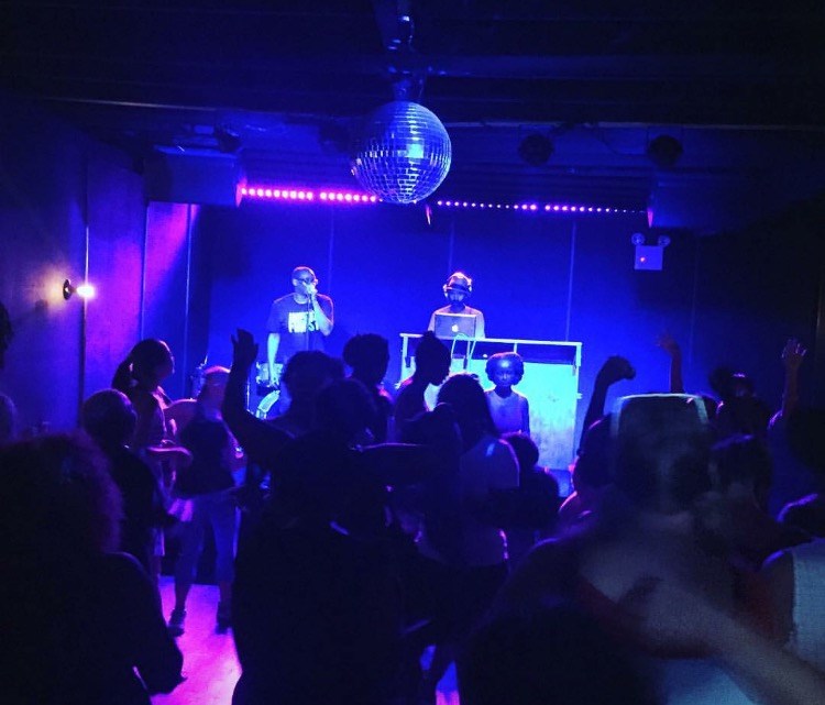 People dance to music in a room lit with blue and purple. A dj plays music in the back.