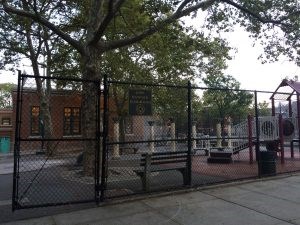 A playground on the other side of a fence. A tree grows in the middle of several benches and columns. A sign labels the park "John Hancock Playground."