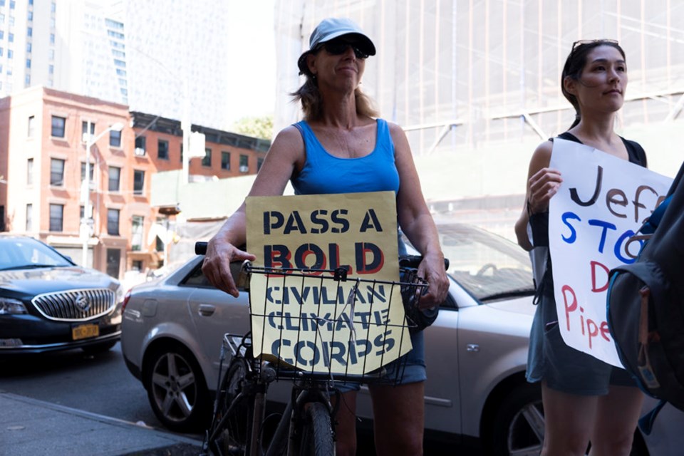 A woman stands behind the basket of her bicycle, in which is a yellow sign that reads "Pass a bold civilian climate corps."