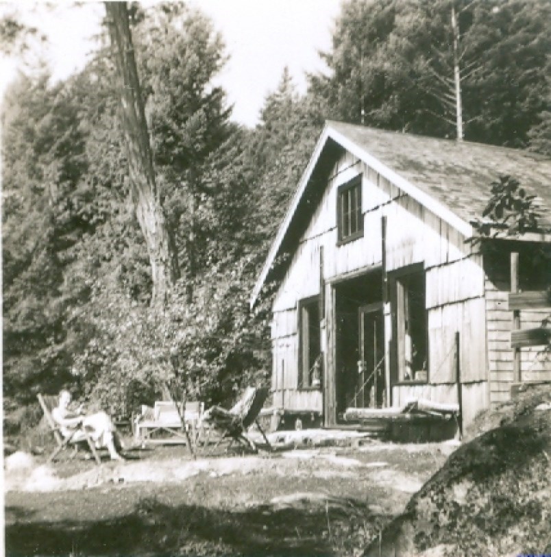 Black and white photograph of two people sitting outside of a barn-looking building in the woods