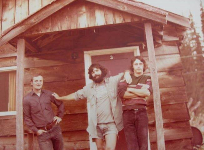 Three men standing in front of a rustic-looking cabin in 1970s