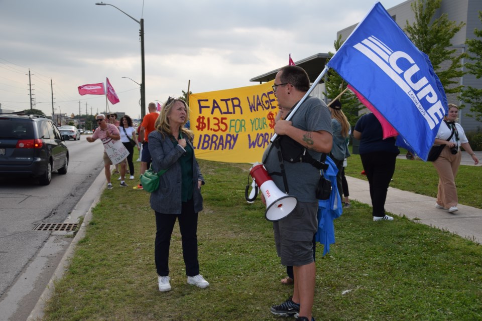 In the foreground at left, Marit Stiles, leader of the Ontario New Democratic Party and the Official Opposition, speaks with a library supporter on the picket line outside the Bradford West Gwillimbury Public Library on Wednesday evening, Aug. 2. In the background at left, Fred Hahn, president of the Canadian Union of Public Employees (CUPE) Ontario rallies support from people driving along Holland Street West.