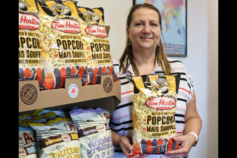 Owner of From Farm to Table Canada, Becky Smollett, poses by a shelf of different popcorn brands holding a bag of butter and salt flavoured popcorn.