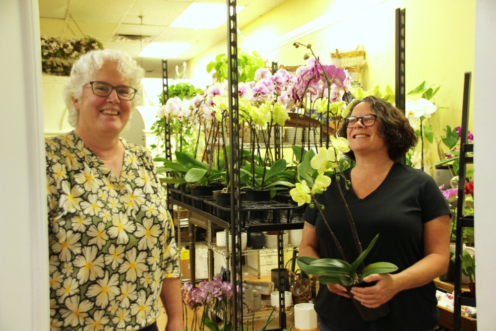 Two smiling women stand next to a large refridgerator of flowers, while one holds a bouquet. 