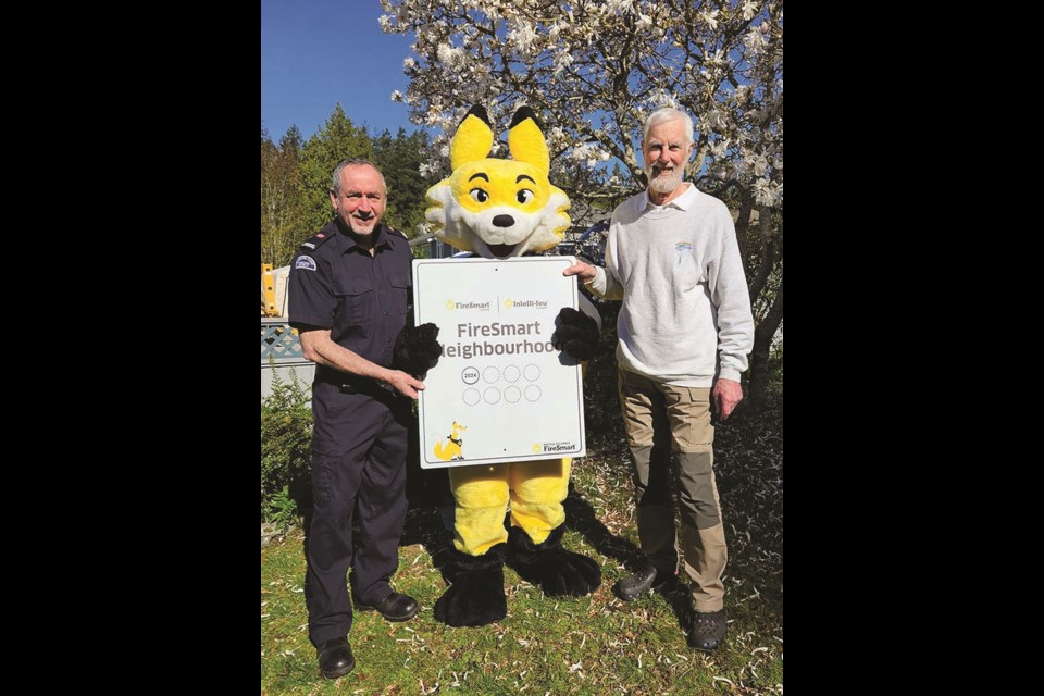 Woodcreek Park’s “FireSmart Certification” plaque is held up by FireSmart coordinator David MacIlwraith, FireSmart mascot “Ember,” and resident Buzz Bennett.