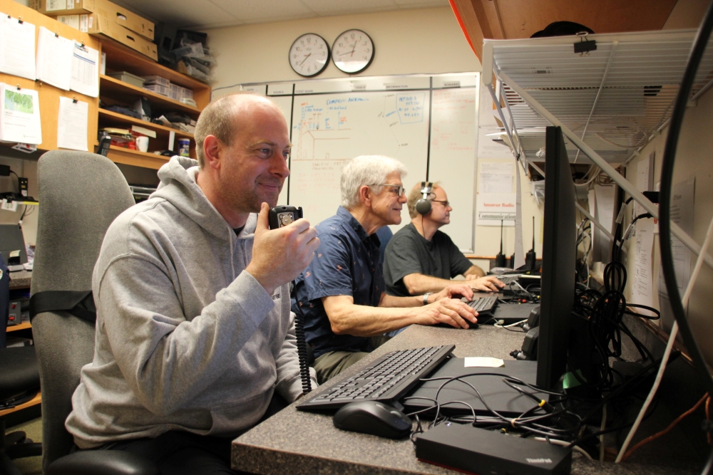 Three men sit at computers, with hand radios, demonstrating radio communications.