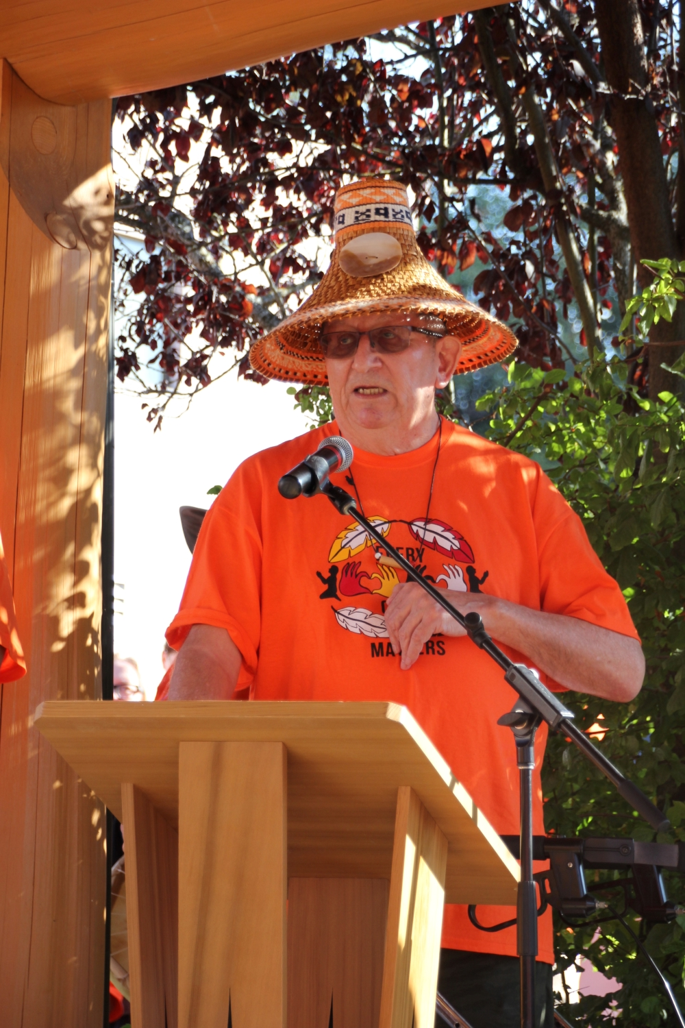 Garry Feschuk stands behind a podium, wearing orange and a woven cedar hat.
