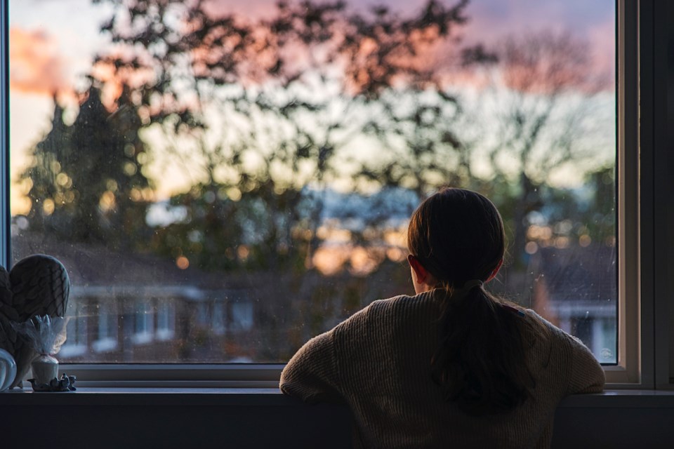 A young girl, with a ponytail, looks out of her bedroom window as the sun is setting.