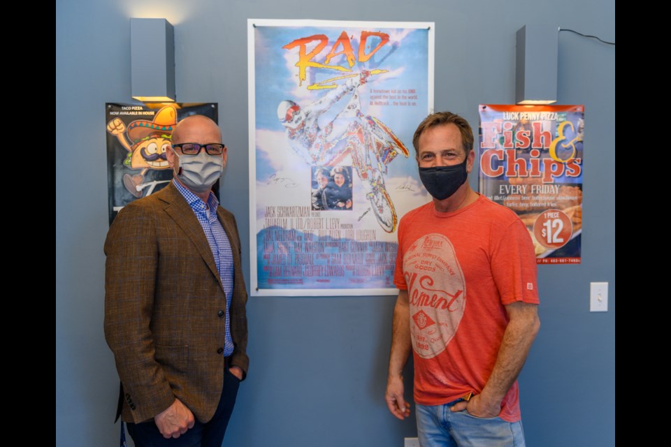 Mayor Jeff Genung and Tim Hall of Lucky Penny Pizza pose for their photo beside the signed poster for the movie RAD at Lucky Penny Pizza in Cochrane on Tuesday (April 13). (Photo by Jack Carter/The Cochrane Eagle)