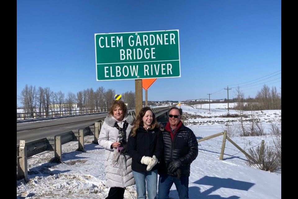 Clem Gardner’s relatives at the sign unveiling. L-R: Janet Hawes (widow of Mike Hawes, grandson), Haley Hawes (great, great granddaughter) and Kevin Hawes (great grandson).
Alberta Transportation placed new signs on the bridge over the Elbow River on Highway 22 on March 7. The bridge was originally dedicated to Clem Gardner for his work with the Canadian Army back in the 1940s when they erected a log bridge as a training exercise and improved community access, presumably including access to then Sarcee Barracks. A commemorative plaque was placed honouring the Clem Gardner name on the modern bridge when it replaced the log structure. The bridge has since been renovated and the plaque was misplaced or possibly grew legs. An ad hoc volunteer group has raised the need for identification of the bridge name especially its place on ‘The Cowboy Trail’. Clem Gardner is still a much honoured and respected cowboy who ranched many years in the nearby Pirmez Creek and Highways 17/22 area. He is well-celebrated especially in a feature statue on the Calgary Stampede Grounds.