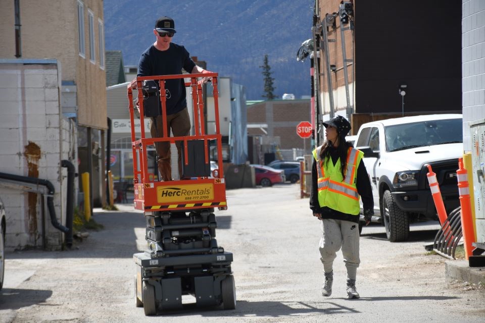 UpLift! co-creator Oliver Andrew drives a lift with Argentinian muralist Fio Silva walking alongside in Jasper, Alta. on Thursday, May 1, 2025. Silva created a phoenix mural behind Whistlers Inn for the UpLift! Jasper Mural Festival. Silva created a phoenix mural for the UpLift! Jasper Mural Festival.