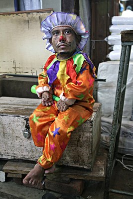 A "Little Person" working as a clown in a textile bazaar in Mumbai.
Photographed while on a break, the man spends each day dancing for customers in front of his employer's stall in order to attract business.
