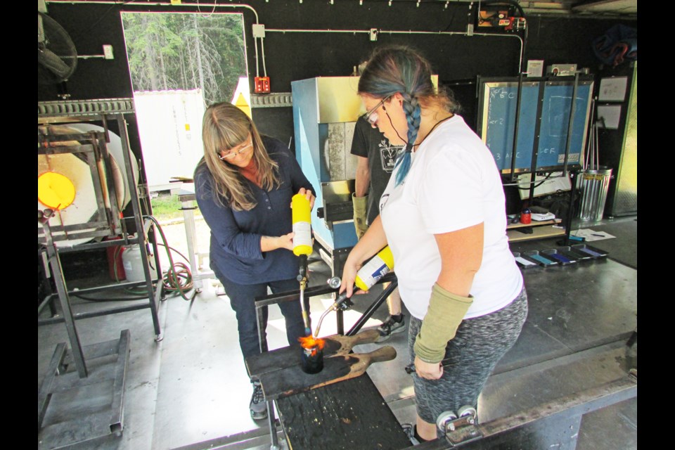 New glass blower Denise Wasnik and instructor Mandy Patchin of Glass House Xperience, a mobile glass blowing facility, put fire to the bottom of the newly-formed drinking glass to get rid of the nub where the glass was held to the pipe during its creation.