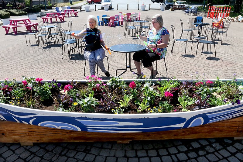 MARIO BARTEL/THE TRI-CITY NEWS Coun. Zoe Royer, the chair of Port Moody's arts and culture committee, and Brenda Millar, who's leading the effort of the committee to enliven the Queens Street plaza, make bubbles from bubble-makers stored in one of two treasure chests filled with kids activities that are now installed at the plaza, along with brightly-painted chairs and tables, and a garden planted in a canoe decorated with symbols from First Nations culture.