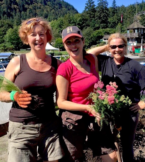 Three women smiling at the base of trunk rd. Two are holding plants.