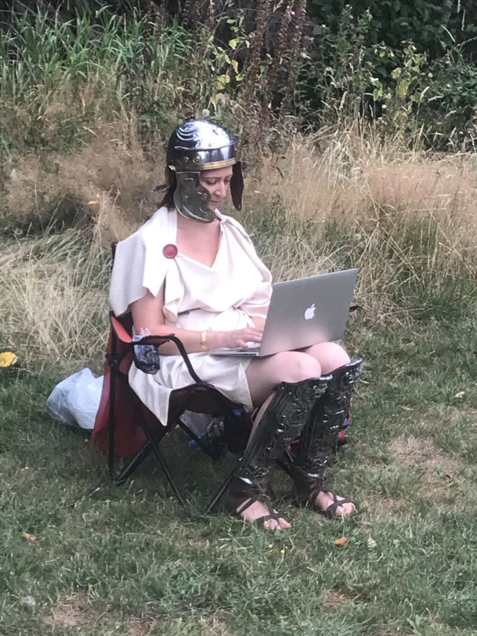 Woman in a winged armoured helmet and Grecian costume sitting in a chair, typing on a mabook