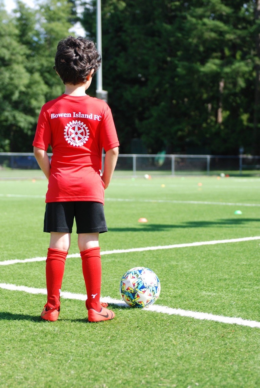 Young child in soccer uniform standing on turf with back to camera and soccer ball in front of them