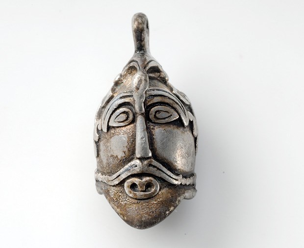 Pendant in the shape of a male head. On the head there is a bird with its wings spread out on the side down to the ears. The pendant comes from a unique grave find containing objects of precious metals, marking the deceased as of great importance in society. The grave belonged to a woman in Aska, Hagebyhöga, Östergötland, Sweden.