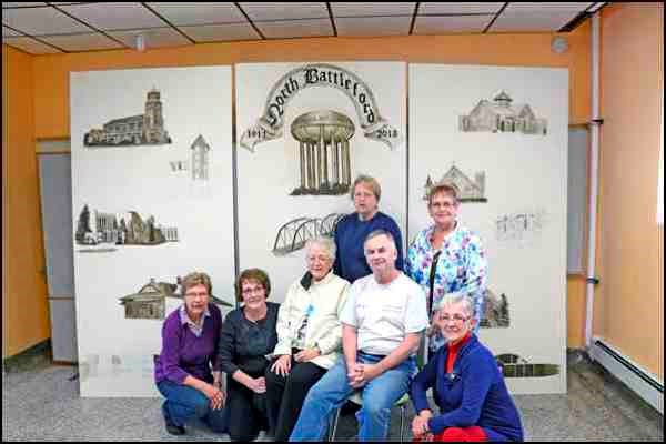 Members of the Battlefords Art Club in front of a partially completed mural that will be used during the centennial celebrations to take place on New Year&#8217;s eve. The mural depicts a variety of local landmarks, which were drawn, then projected onto the painting surface to enlarge them. After tracing the projections, the art club began the process of painting in sepia, a new process for some of the members.