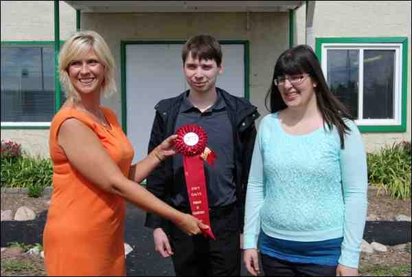 A first place ribbon in the non-commercial category of the North Battleford 100th anniversary Northwest Territorial Days Parade was a pleasant and welcome surprise for Prairie Employment Program summer students Sarah Leitner (right) and Zack Startup (centre). Born, raised and educated in the Battlefords, Leitner and Startup are pursuing university programs to become elementary or secondary school teachers. They were employed this summer as assistant job counsellors with Prairie Employment and, as part of their summer work experience, designed and helped to build this year&#8217;s award winning parade entry. Presenting the red ribbon is Territorial Days parade sponsor, Battlefords&#8217; Agricultural Society Executive Director Jocelyn Ritchie (left).