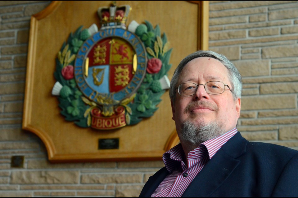 Local graphic designer and heraldic artist Allan Ailo stands in the foyer of New Westminster city hall by a colourful example of local heraldry, the badge of the Royal Engineers.
