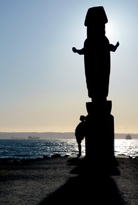 A passerby enjoying the view leaning up against the Guardian Totem Coast Salish Totem welcome figure on a windy Autumn day at Ambleside.