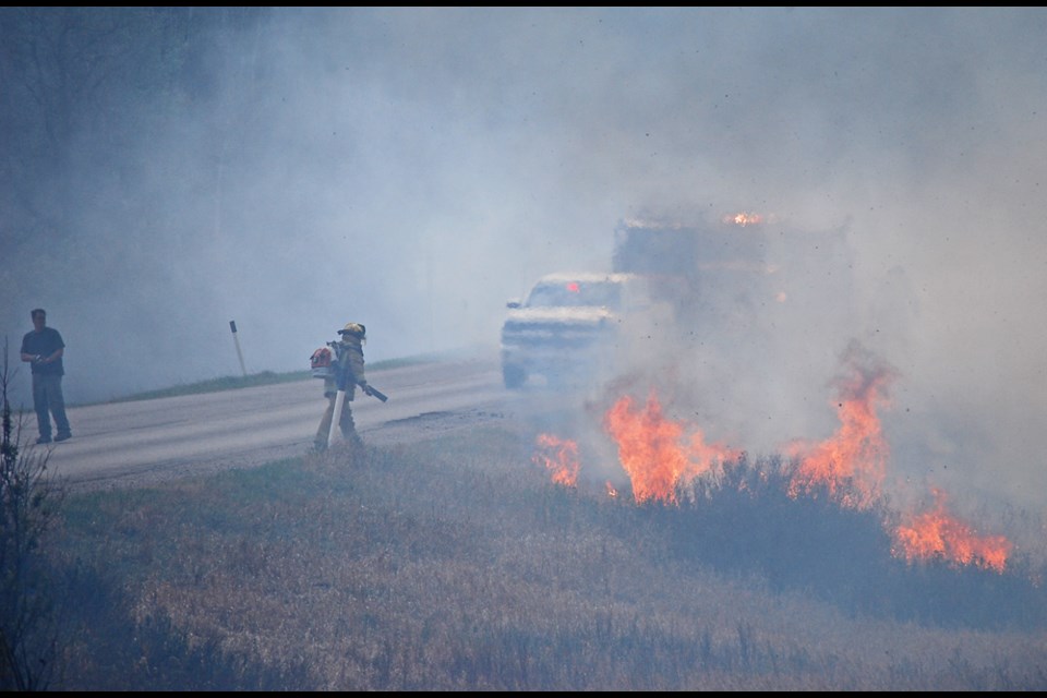 With billowing smoke on PR 259 in the Assiniboine Valley about supper time on Tuesday, May 15, a man is helping traffic to safely navigate up the valley hill. Firefighters are focused on their job fighting fire on the steep grade of the road that drops off into the valley ravine below.