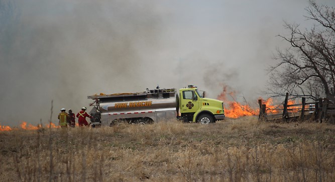 Wallace District Fire Department at a grass fire southeast of Virden, just about three kms from town today, Thursday, April 24 at about 1:30 p.m. High wind is fanning flames as firefighters hose down the leading edge of a fire that was raging in the long grass of an old farm yard. Flames engaged the aged wooden fence posts in the fire’s path.