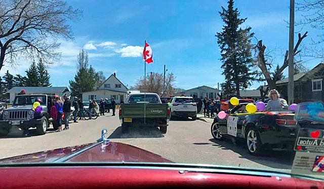 A picture from the passenger seat of a vintage two-door hardtop (owned/driven by Gordon White), shows a car ahead to the right decorated with balloons and a sign - a message from family and friends to a senior during the COVID-19 shut-in. Looking further down the street, a green truck is driven by Lloyd and Carol Williams and just ahead, (not visible) Kevin Williams leads the motorcade in a 52 Fargo originally owned by Jack Chapman, now himself a resident in Evergreen Place in Virden.