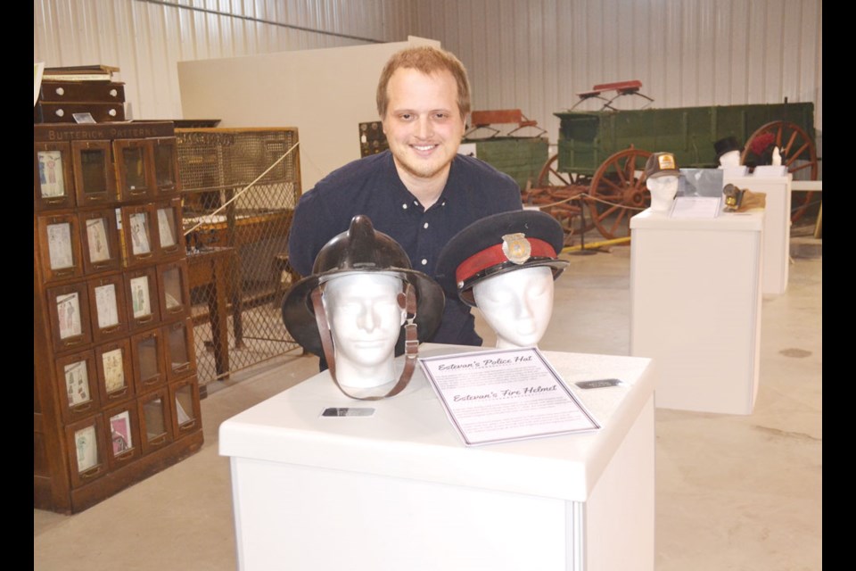 Souris Valley Museum curator/director Mark Veneziano crouches between hats for police and the fire department.