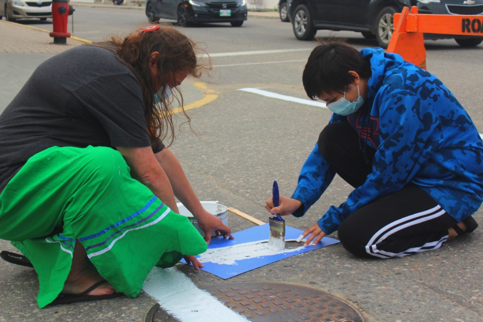Loretta McDermott and Trina Bear paint the All Children Matter crosswalk on Flin Flon’s Main Street June 19. The crosswalk, which features white feathers and large “All Children Matter” text, pays tribute to Indigenous children and survivors and victims of the Canadian residential school system. - PHOTO BY ERIC WESTHAVER
