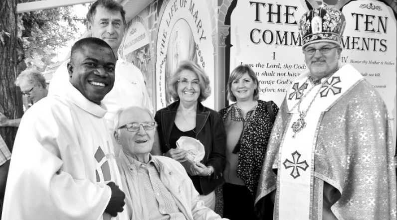 Rev. Franklin Emereuwa of Canora (left) and Bishop Bryan Bayda (right) joined Cas and Marie Broda of Kamsack and their daughter Shannon (Brad) Vanin in front of images which created a mural on the Parish Hall wall.