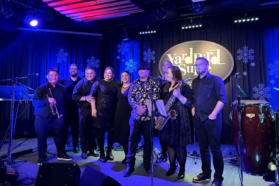A group of nine jazz musicians stand smiling on stage at Edmonton’s Yardbird Suite under blue and pink lights. / Yardbird Suite | Facebook