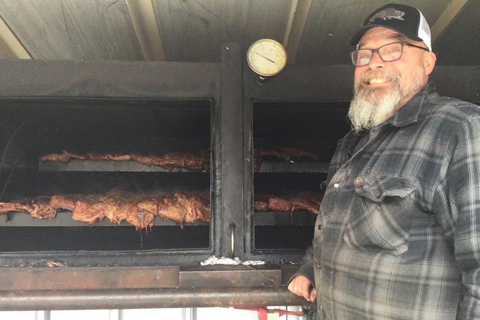 A man stands smiling in front of a large smoker filled with delicious meat at Twin Butte Country General Store. /Twin Butte Country General Store | Facebook
