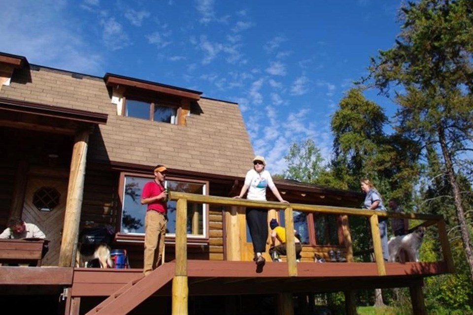 A group of three friends stand on a patio of a log cabin at Forest House in Saskatchewan with trees all around. Tourism Saskatchewan.