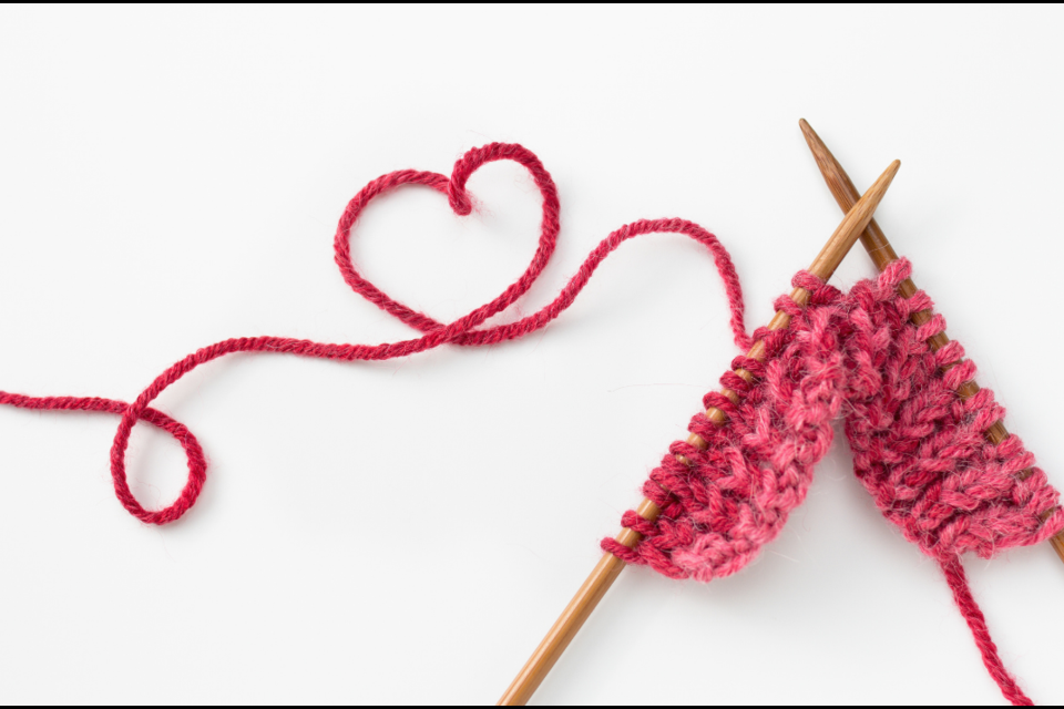 Knitting needles with part of a yarn project on them. Extending from the project is pink yarn in the shape of a heart.