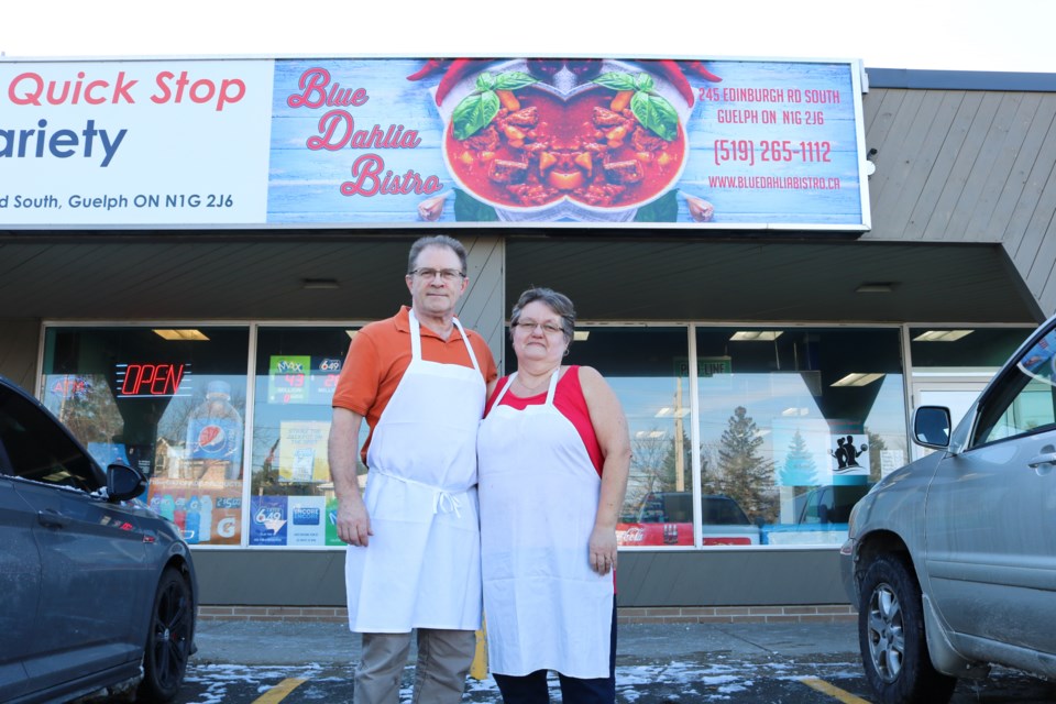 Mike Durcik and Sylvia Durcik posing below the sign for Blue Dahlia Bistro, a takeout restaurant specializing in Hungarian and Central European food.