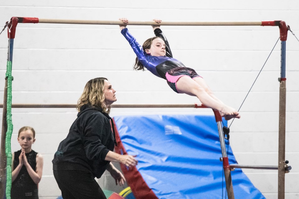 A gymnast shows off her skills on the bars with a coach watching nearby.