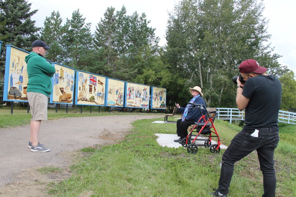 Elk Point Historical Mural artist Billie Milholland tells Brian Siddle of Strong Coffee Marketing one of the many stories of people and places on the giant artwork while Siddle’s cameraman snaps a photo of the artist. Vicki Brooker photo.