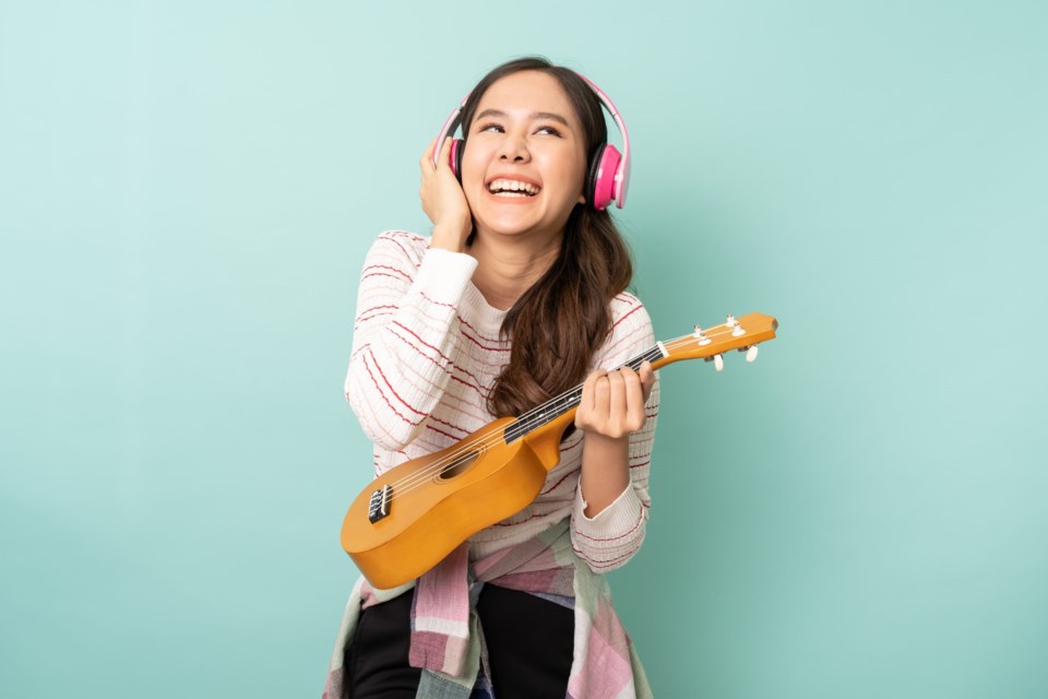 Young,Asian,Woman,Using,Headphones,While,Playing,Ukulele,With,Happy.
