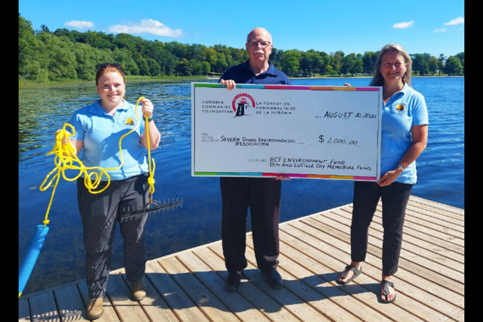 HCF Executive Director, Scott Warnock presents cheque to SSEA Executive Director, Julie Cayley while Emily Edgley, SSEA’s Citizen Science and Water Quality Coordinator (left), holds one of the aquatic plant rakes purchased.