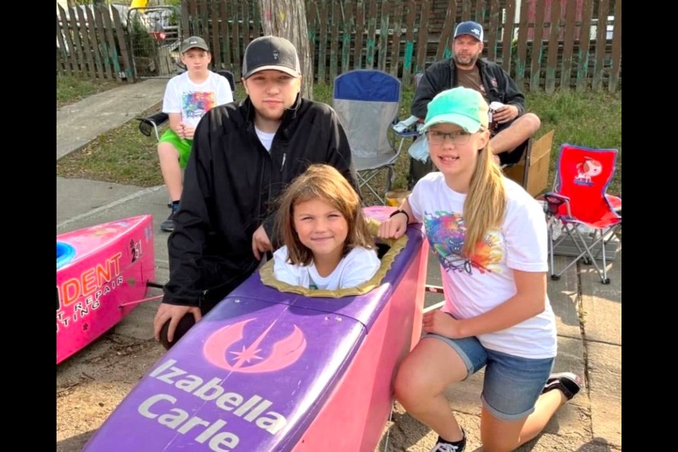 Nine-year-old Izabella Carle sits in her soapbox race car, proudly bearing her name. The car has a long family history, previously raced by her uncle Jordan Carle (2009–2011), left, and cousin Danica Finlay (2017–2019), right.