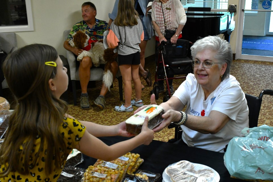 Resident Dee Wilby (right) hands a freshly purchased baked good to a young patron (left) during her bake sale at the Atria Mulberry Estates grand reopening event.