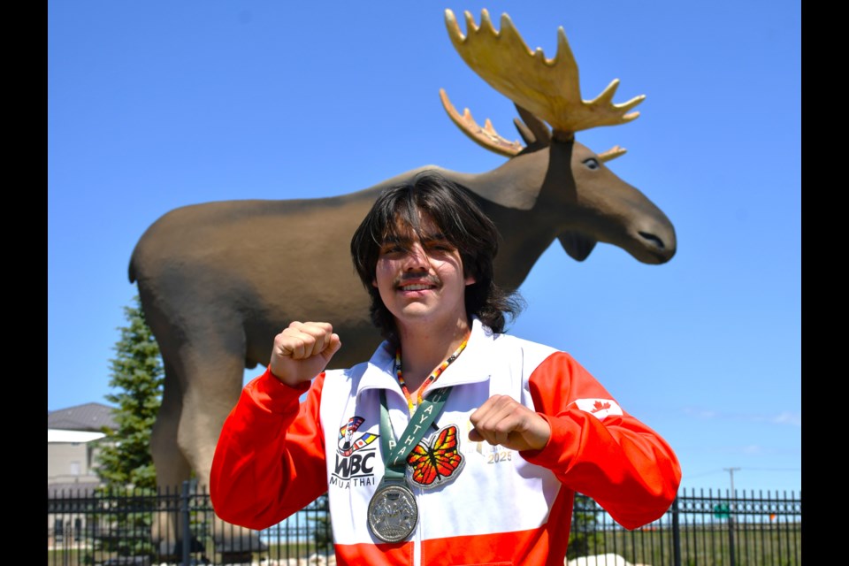 Cameron Billy stands in front of Mac the Moose wearing his silver medal and traditional beaded butterfly medallion — symbols of achievement and transformation after representing Moose Jaw on the world stage.