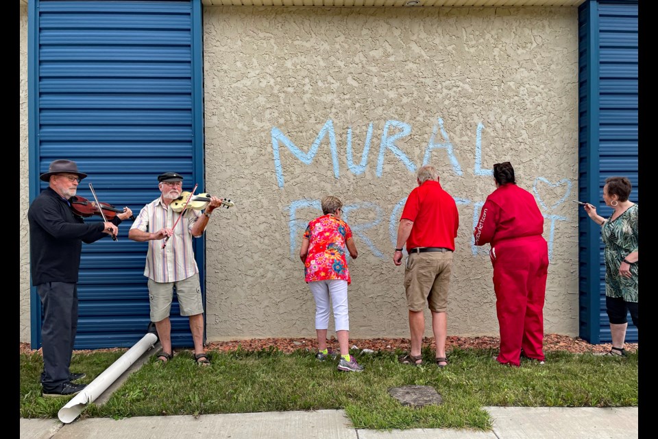 With uplifing fiddle music providing inspiration, local artist Karen Scarlett (second from right) leads volunteers in the start of the ambitious mural project on the south wall of the Innisfail Royal Canadian Legion Branch #104 during the late afternoon of July 10. The public art project is expected to take Scarlett and dozens of volunteers two months to complete. Johnnie Bachusky/MVP Staff