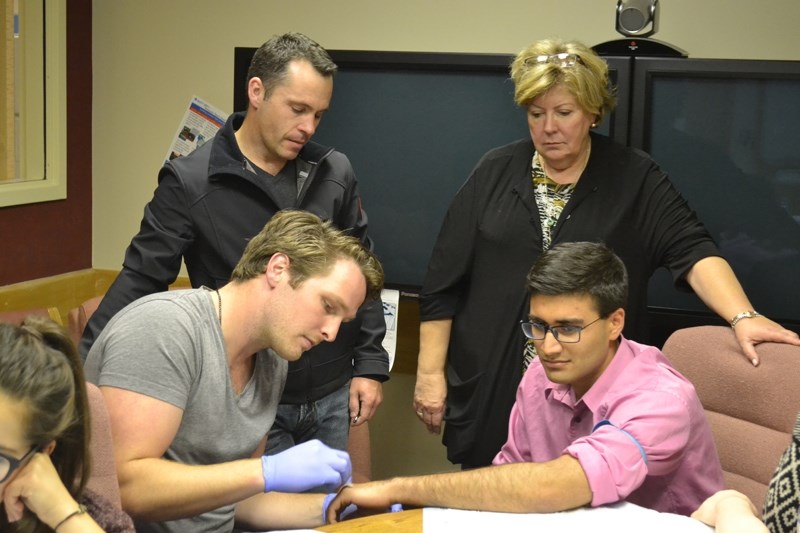 A physio student prepares to start an IV on Arth Pahwa&#8217;s arm as instructor Dr. Jaco Hoffman and Rosemary Burness of the Rural Physician Action Plan look on.