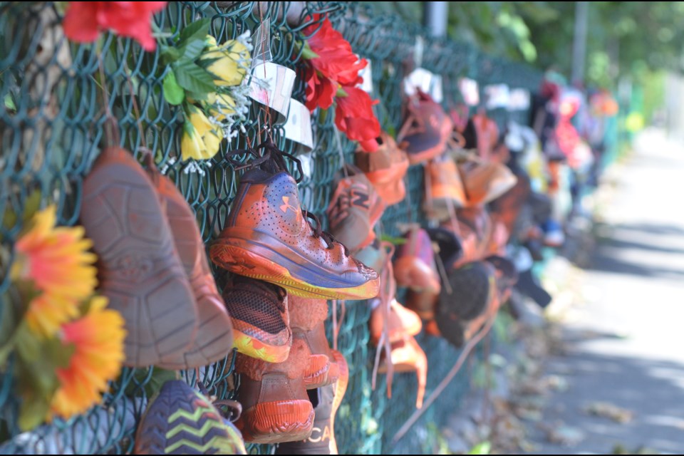 Orange shoes line a chain-link fence adjacent to the sidewalk on the north side of Lynn Valley Road, just east of Pioneer Square, created by Gwynneth Powell Sobejko with the help of the community. 