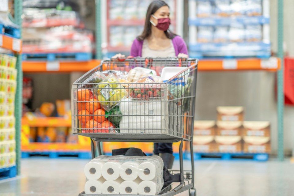 A young female adult is pushing her shopping cart full of groceries down an aisle at a supermarket warehouse. 