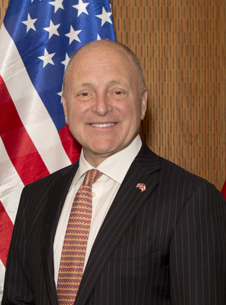 Older balding man in suit in front of the US Flag | US State Department